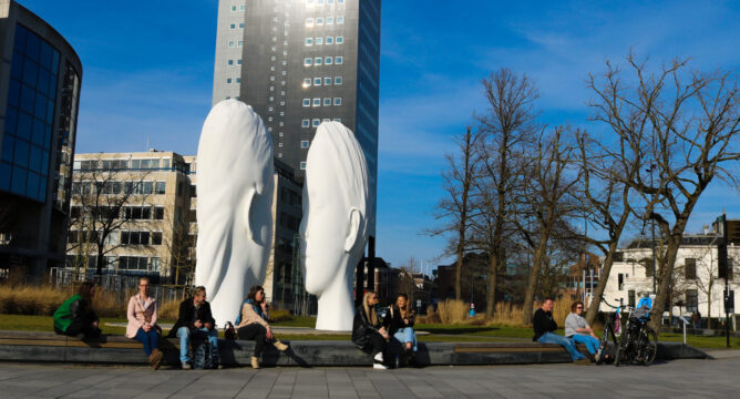 Beeld van de twee hoofden bij het station in Leeuwarden met op de achtergrond de Achmea Toren en op de voorgrond mensen die op de rand langs het trottoir zitten